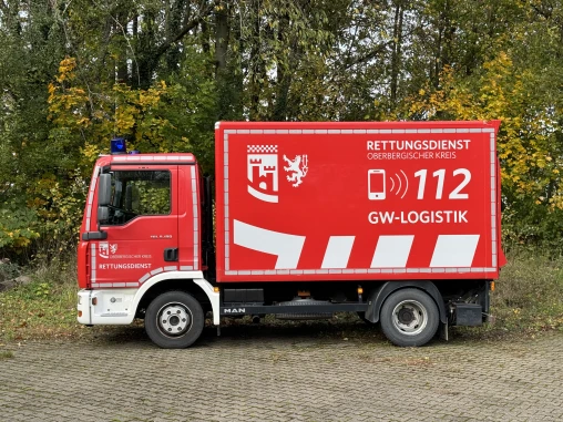 Ein rotes Einsatzfahrzeug mit der Aufschrift 'Rettungsdienst Oberbergischer Kreis' und 'GW-Logistik'. Das Fahrzeug steht auf einem gepflasterten Untergrund vor Bäumen.