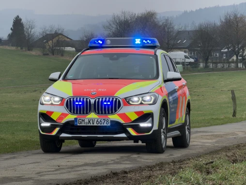 Ein Einsatzfahrzeug der Rettungsdienste, ein BMW SUV mit rot-gelber Warnlackierung und eingeschaltetem Blaulicht, steht auf einem ländlichen Weg. Im Hintergrund sind eine Wiese, Bäume und Häuser sichtbar.