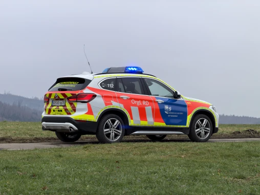 Ein Einsatzfahrzeug des Rettungsdienstes, ein BMW SUV mit auffälliger rot-gelber Lackierung und Blaulicht, steht auf einer Landstraße neben einer Wiese. Im Hintergrund sind Hügel und bewölkter Himmel sichtbar.