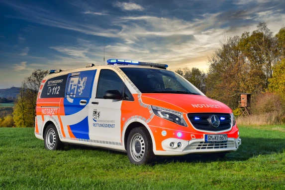 Ein Notarztfahrzeug des Rettungsdienstes Oberbergischer Kreis, Mercedes-Benz, mit orange-weißer und blauer Lackierung, steht auf einer Wiese. Im Hintergrund sind Bäume und ein Hochsitz sichtbar.