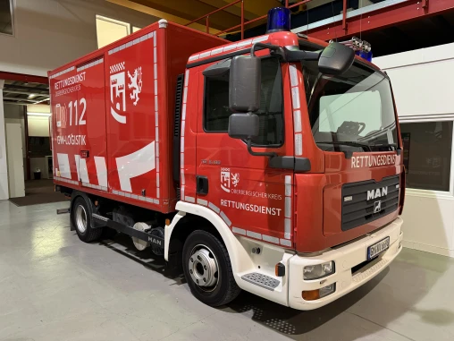 Ein rotes Einsatzfahrzeug des Rettungsdienstes mit der Aufschrift 'Rettungsdienst Oberbergischer Kreis GW-Logistik 112' steht in einer Halle. Das Fahrzeug ist ein MAN-Lkw mit Blaulicht auf dem Dach.