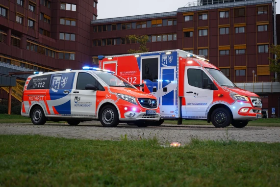 Zwei Rettungsfahrzeuge mit auffälliger rot-weißer Beschriftung und aktivem Blaulicht stehen vor einem mehrstöckigen Gebäude. Im Vordergrund ist Gras sichtbar.