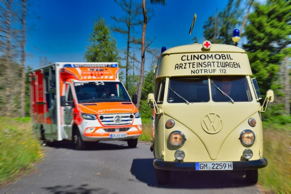 Ein beige Volkswagen-Transporter mit der Aufschrift 'Clinomobil Arzteinsatzwagen Notruf 112' fährt vor einem modernen orange-weißen Rettungswagen auf einer Straße. Im Hintergrund sind Bäume sichtbar.