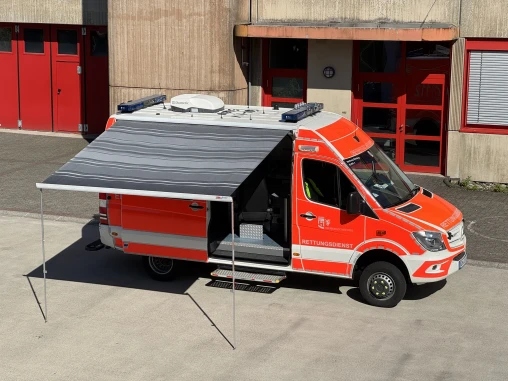 Ein orange-weißes Rettungsfahrzeug mit ausgefahrener Markise steht vor einem Gebäude mit roten Türen. Die Seitentür des Fahrzeugs ist geöffnet.