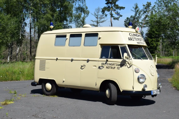 Ein cremefarbenes historisches Rettungsfahrzeug, vermutlich ein VW-Bus, mit der Beschriftung 'Arzteinsatzwagen Notruf 112' und Blaulicht auf dem Dach. Das Fahrzeug steht auf einer asphaltierten Fläche, umgeben von Bäumen und Gras.