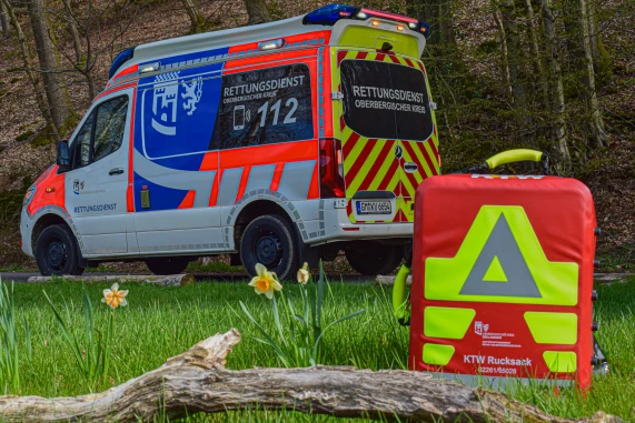Ein Rettungswagen des Oberbergischen Kreises steht in einem Waldgebiet. Im Vordergrund ist ein roter Notfallrucksack mit gelben und reflektierenden Markierungen auf einer Wiese zu sehen, umgeben von Blumen und einem Holzstück.