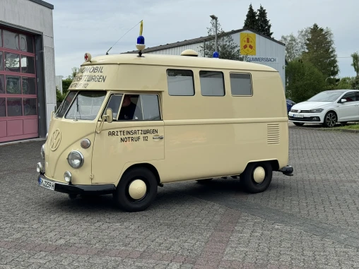 Ein cremefarbener Oldtimer-Krankenwagen mit der Aufschrift 'Arzteinsatzwagen Notruf 112' und einem blauen Licht auf dem Dach, geparkt vor einem Gebäude.