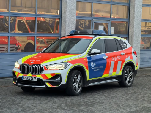 Ein Einsatzfahrzeug des Rettungsdienstes, ein BMW-SUV mit auffälliger rot-gelber Markierung, steht vor einer Fahrzeughalle mit geschlossenen Garagentoren.