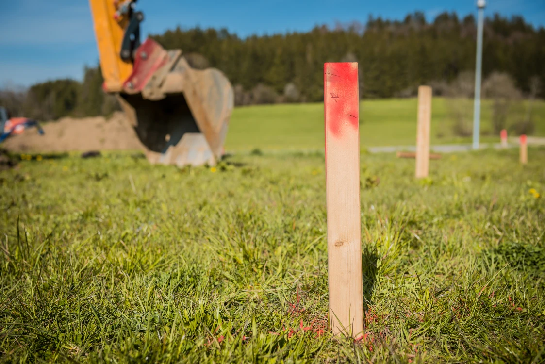 Holzpfähle mit roter Markierung stehen auf einer grünen Wiese. Im Hintergrund ist eine Baggerschaufel und eine Landschaft mit Bäumen sichtbar.
