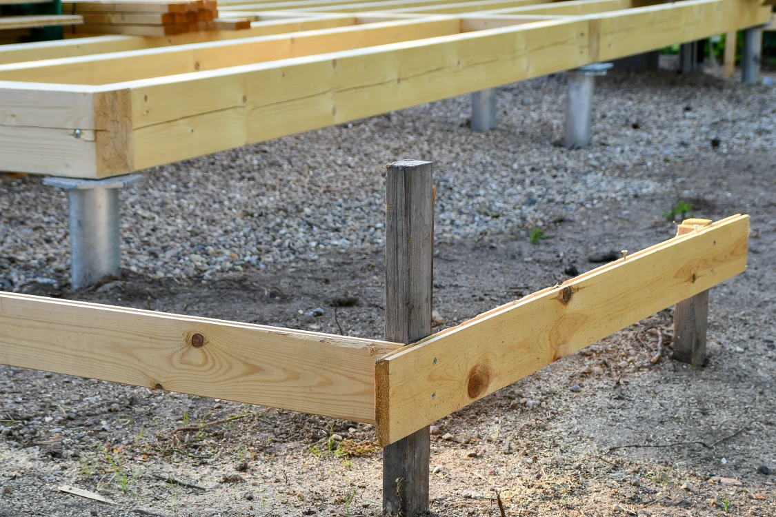 Holzrahmenkonstruktion auf Metall- und Holzpfosten, aufgebaut auf einem mit Kies und Erde bedeckten Untergrund.