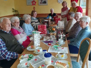 Eine Gruppe älterer Menschen sitzt an einem festlich gedeckten Tisch mit weihnachtlicher Dekoration, Geschenken und Kaffee. Zwei Personen in Nikolauskostümen stehen dahinter.
