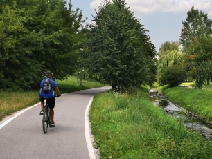Ein Radfahrer mit Helm und Rucksack fährt auf einem asphaltierten Weg durch eine grüne Landschaft. Rechts neben dem Weg fließt ein schmaler Bach.