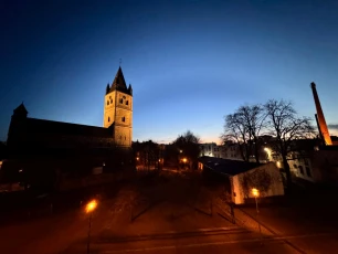 Abendliche Stadtansicht mit einer beleuchteten Kirche links und einem hohen Schornstein rechts. Dazwischen Bäume, Straßenlaternen und Gebäude.