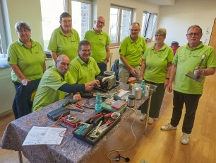 Eine Gruppe von acht Personen in grünen Polohemden steht und sitzt um einen Tisch mit Werkzeugen und Elektrogeräten in einem Raum mit Fenstern.