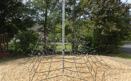 Ein Klettergerüst in Form eines Netzes, das an einem zentralen Metallpfosten befestigt ist, steht auf einem mit Holzschnitzeln bedeckten Boden. Im Hintergrund sind Bäume und Büsche zu sehen.