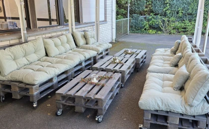 Eine Sitzgruppe aus Holzpaletten mit hellgrünen Polstern, bestehend aus zwei Sofas und zwei Tischen. Die Tische sind mit Kerzen und Dekoration verziert. Im Hintergrund ist eine Hauswand und eine Hecke zu sehen.