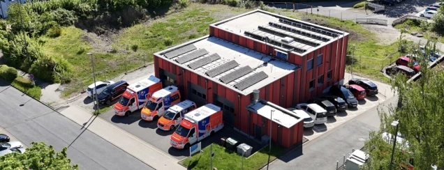 Ein rotes Gebäude mit Solarpanelen auf dem Dach, davor mehrere Rettungswagen und geparkte Autos. Das Gelände ist von Grünflächen und einem Zaun umgeben.
