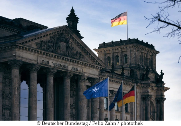 Das Reichstagsgebäude in Berlin mit dem Schriftzug 'Dem Deutschen Volke' auf dem Giebel. Im Vordergrund wehen die Flaggen der NATO, EU und Deutschland.