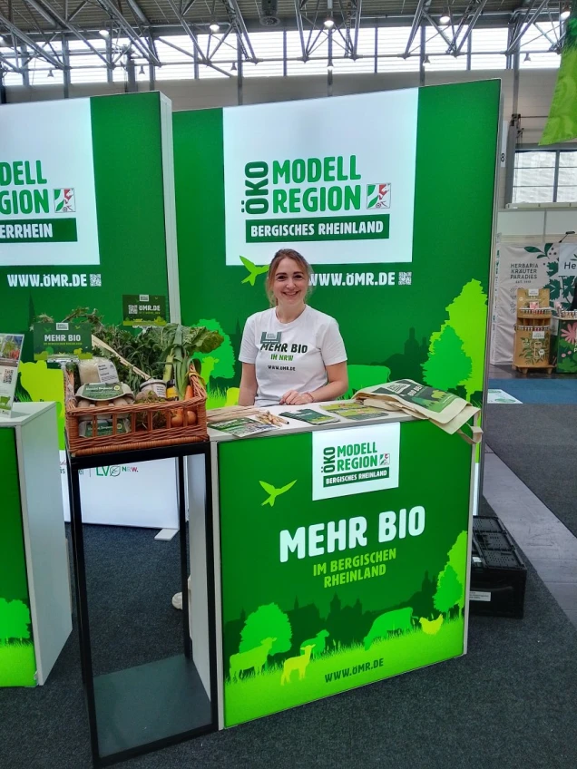 Eine Frau steht hinter einem Messestand der Öko-Modellregion Bergisches Rheinland. Der Stand ist grün gestaltet und zeigt den Slogan 'Mehr Bio im Bergischen Rheinland'.
