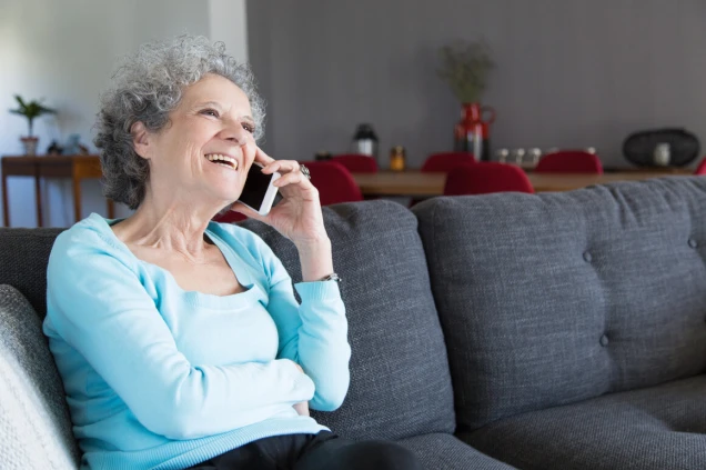 Ältere Frau mit lockigem grauem Haar sitzt lächelnd auf einem grauen Sofa und telefoniert mit einem Smartphone. Im Hintergrund ist ein Essbereich erkennbar.