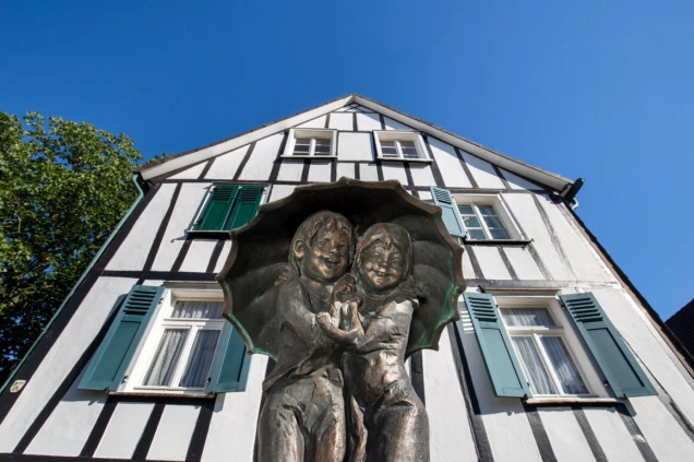 Bronzestatue von zwei Kindern unter einem Regenschirm vor einem Fachwerkhaus mit grün-blauen Fensterläden und weißer Fassade unter blauem Himmel.