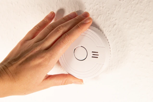 Eine Hand berührt einen an der Decke montierten weißen Rauchmelder mit rundem Design und Lüftungsschlitzen.