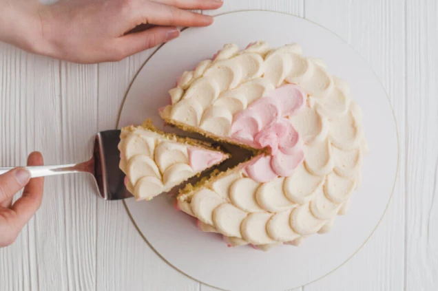 Eine Person schneidet ein Stück aus einer rosa-weißen Torte mit Wellenmusterglasur heraus. Die Torte liegt auf einem weißen Untergrund.