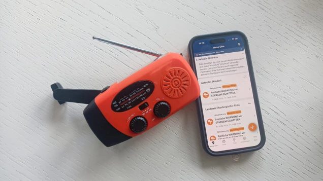 Ein orange-schwarzes Kurbelradio mit ausziehbarer Antenne liegt neben einem Smartphone auf einer weißen Oberfläche. Auf dem Smartphone ist eine Wetterwarnungs-App geöffnet.