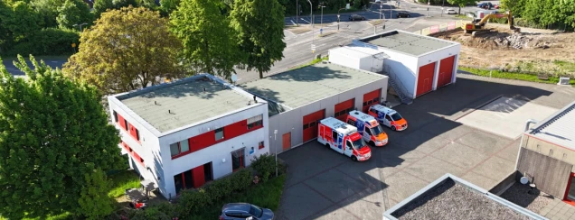 Luftaufnahme einer Rettungswache mit zwei Gebäuden und mehreren roten Garagentoren. Drei Rettungswagen stehen davor. Im Hintergrund sind Bäume und eine Baustelle sichtbar.
