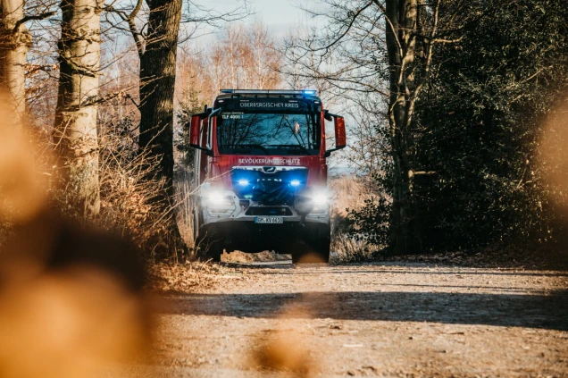 Ein rotes Feuerwehrfahrzeug mit eingeschalteten Blaulichtern fährt auf einem Waldweg. Bäume und trockenes Laub umgeben den Weg.