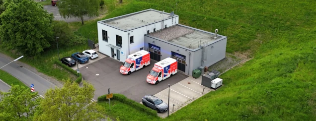 Luftaufnahme einer Rettungswache mit zwei Rettungswagen vor den Garagentoren. Das Gebäude ist von Grünflächen und einigen parkenden Autos umgeben.