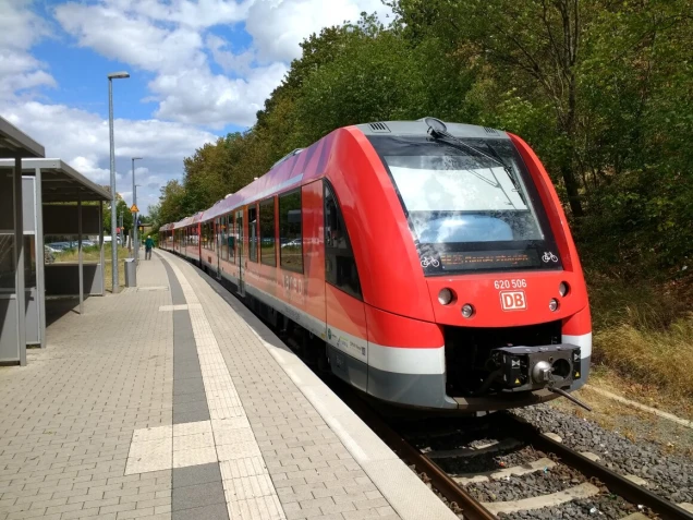 Ein roter Regionalzug der Deutschen Bahn mit der Aufschrift 'RB 25' steht an einem Bahnsteig. Der Bahnsteig ist gepflastert und mit einem Wartehäuschen ausgestattet. Im Hintergrund sind Bäume und ein bewölkter Himmel zu sehen.