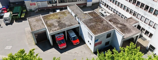 Gebäude mit Flachdach und Moosbewuchs. Zwei Rettungsfahrzeuge in Garagen. Schild mit 'Corona Ambulanz' an der Wand. Parkplätze und Müllcontainer im Hof.