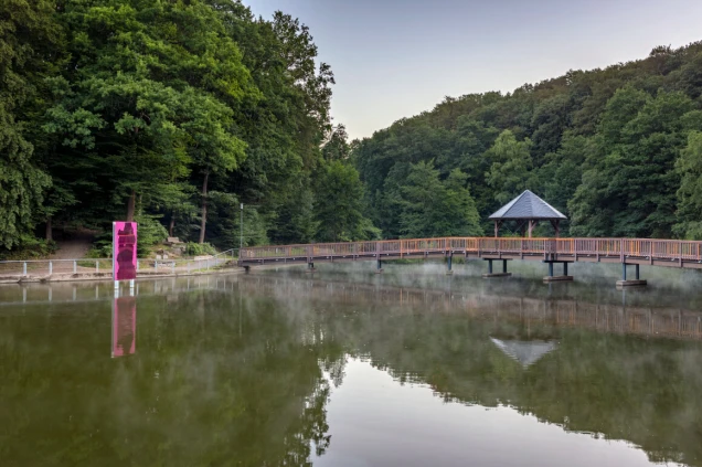 Ein See mit einer Holzbrücke und einem Pavillon, umgeben von dichtem Wald. Im Vordergrund ist ein pinkes Kunstobjekt am Ufer zu sehen.