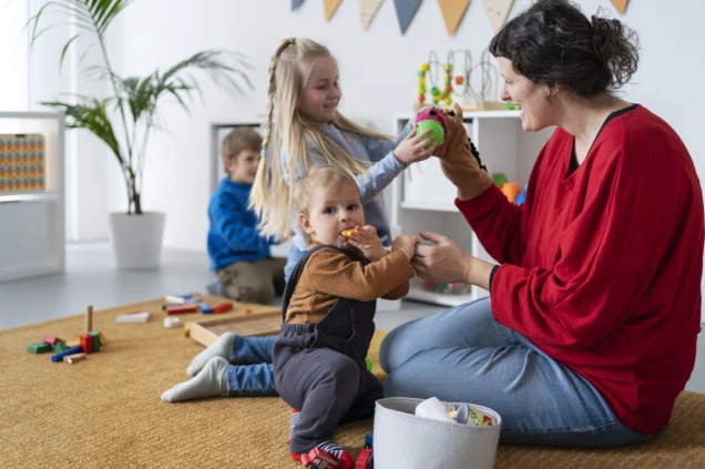 Eine Frau in rotem Oberteil spielt mit drei Kindern in einem hellen Raum. Spielzeug ist auf einem Teppich verteilt, und im Hintergrund stehen Regale mit weiteren Spielsachen.