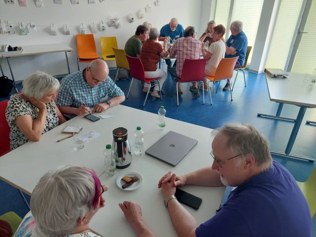 Mehrere ältere Personen sitzen in kleinen Gruppen an Tischen in einem hellen Raum. Auf den Tischen stehen Wasserflaschen, eine Thermoskanne und ein Laptop.