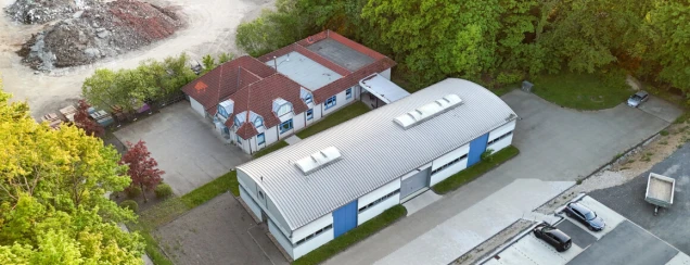 Luftaufnahme eines Gebäudekomplexes mit einem roten Ziegeldach und einer angrenzenden Halle mit gebogenem Metalldach. Links Bäume, rechts ein Parkplatz mit Autos.