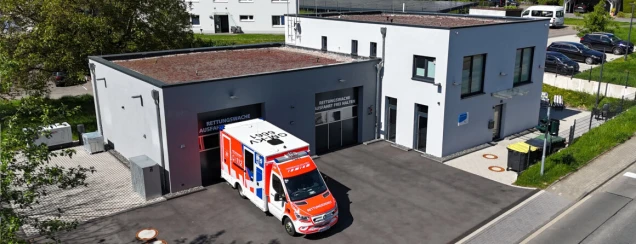 Modernes, einstöckiges Gebäude mit Flachdach und weiß-grauer Fassade. Vor dem Gebäude steht ein Rettungswagen. Im Hintergrund sind geparkte Autos und Grünflächen zu sehen.