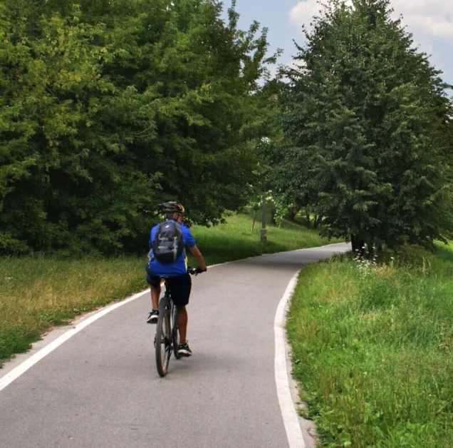 Ein Radfahrer mit Helm und Rucksack fährt auf einem asphaltierten Weg durch eine grüne Landschaft. Rechts neben dem Weg fließt ein schmaler Bach.