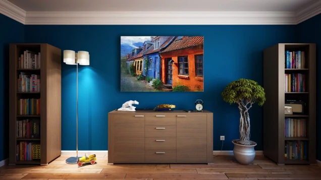 Ein Wohnzimmer mit blauer Wand, zwei Bücherregalen, einer Holzkommode, einer Stehlampe, einem gerahmten Gemälde, einer Zimmerpflanze und Dekoobjekten.