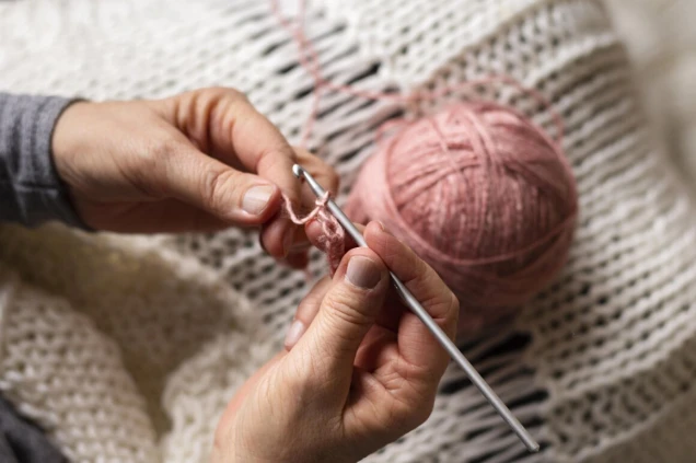 Nahaufnahme von Händen, die mit einer Häkelnadel und rosa Garn häkeln. Im Hintergrund ist ein weißes gestricktes Tuch zu sehen.