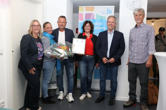 Eine Gruppe von sechs Personen steht lächelnd vor einem Banner mit der Aufschrift 'Herzlich Willkommen'. Eine Frau hält einen Blumenstrauß, eine andere eine Urkunde. Im Hintergrund ist eine Uhr und eine weitere Person erkennbar.