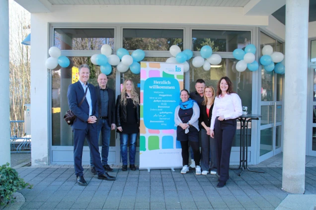 Eine Gruppe von sieben Personen steht vor einem Eingang mit Glastüren, geschmückt mit weißen und blauen Luftballons. Ein Schild mit der Aufschrift 'Herzlich willkommen' in mehreren Sprachen steht in der Mitte.