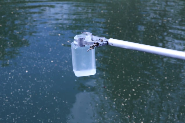 Ein transparenter Behälter mit Wasser wird mit einer metallischen Halterung an einem Stab über eine Wasseroberfläche gehalten. Die Wasseroberfläche ist blau und mit kleinen Partikeln übersät.
