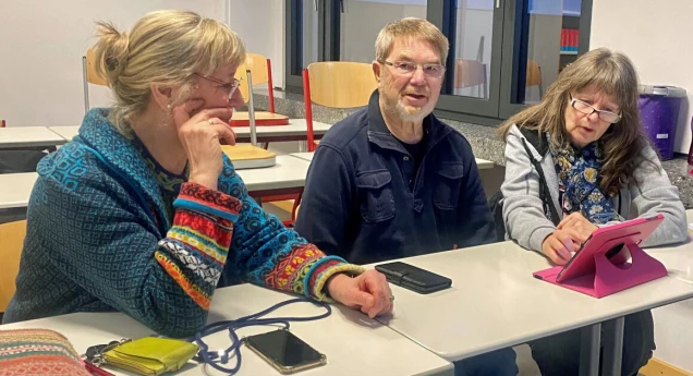 Drei ältere Personen sitzen an einem Tisch in einem Raum mit leeren Stühlen im Hintergrund. Eine Frau benutzt ein Tablet mit pinker Hülle.