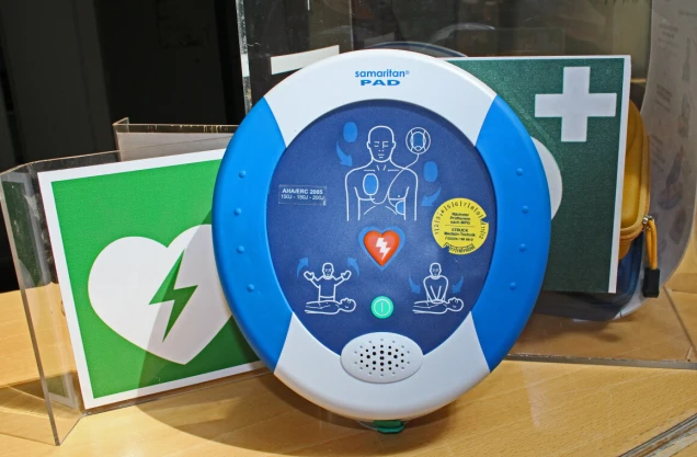 Ein automatisierter externer Defibrillator (AED) der Marke Samaritan PAD auf einem Tisch. Im Hintergrund sind grüne Symbole mit Herz und Kreuz sichtbar.