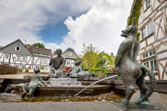 Brunnen mit bronzenen Figuren, darunter Menschen und tierähnliche Gestalten, in einem Platz mit Fachwerkhäusern und Pflanzen. Himmel teils bewölkt.