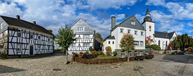 Ein Kopfsteinpflasterplatz mit Fachwerkhäusern, einer Kirche mit Turm und Spitzdach sowie Bäumen im herbstlichen Laub. Im Vordergrund stehen Sitzbänke.