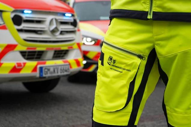Person in gelber Einsatzkleidung des Rettungsdienstes Oberbergischer Kreis steht vor zwei Einsatzfahrzeugen mit eingeschaltetem Blaulicht.