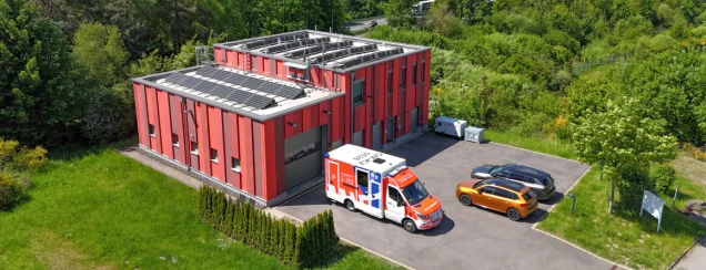Ein modernes, rotes Gebäude mit Solarpaneelen auf dem Dach, umgeben von Bäumen. Auf dem Parkplatz stehen ein Rettungswagen und zwei Autos.